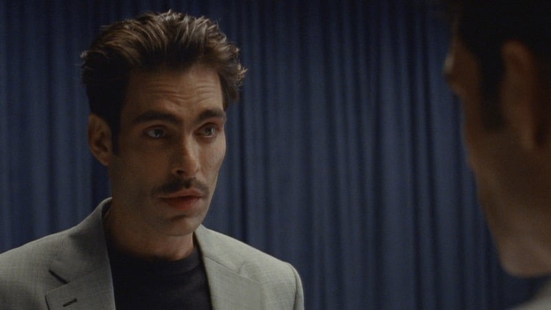 Jon stands in a light gray blazer over a black T-shirt with a mustache, facing another figure in profile against a navy curtain