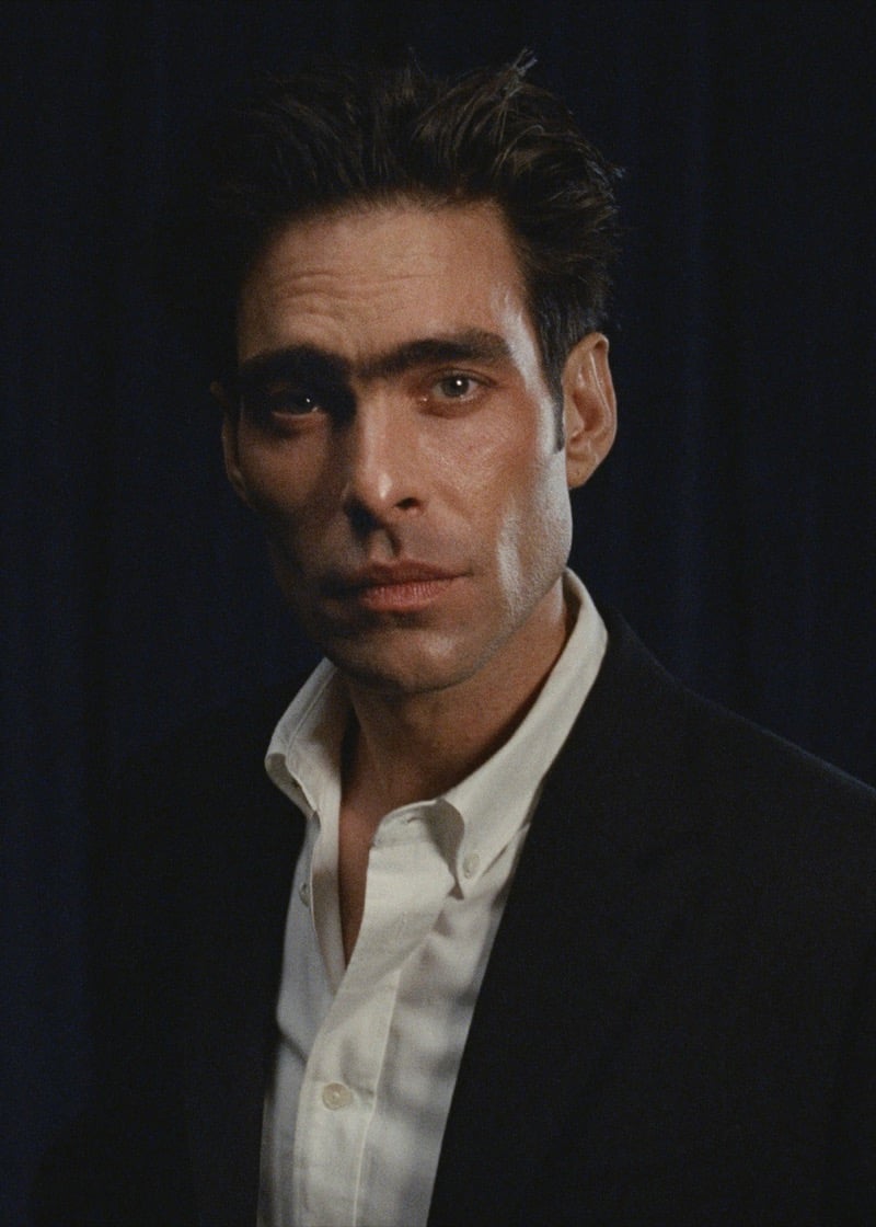Portrait of Jon in a black blazer worn over an open white shirt, against a deep blue curtain