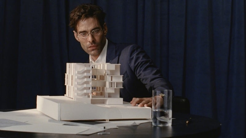 Jon wears round wire-rimmed glasses and a navy suit while studying a white architectural model on a dark table