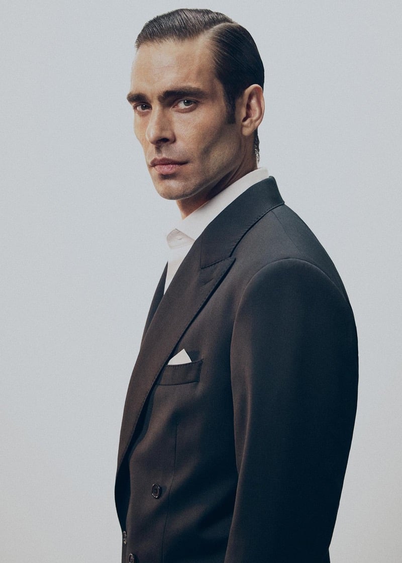 Profile portrait of Jon in a black peak-lapel suit jacket with a white shirt and a white pocket square, hair slicked back