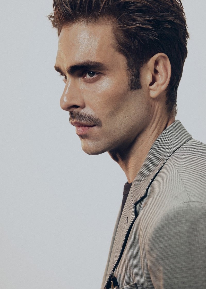Close-up profile of Jon with a mustache, wearing an oat micro-houndstooth blazer over a dark brown T-shirt