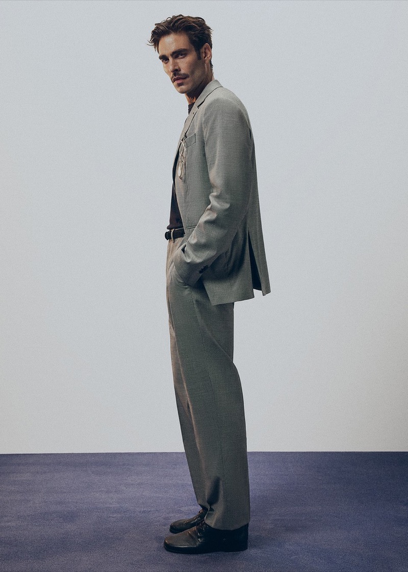 Jon stands in profile in a gray single-breasted suit with a dark T-shirt and chain, brown leather lace-up shoes, on a navy carpet