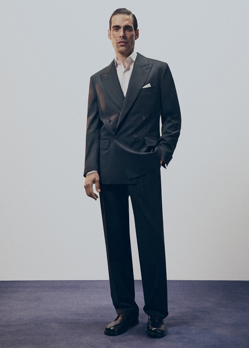 Jon stands in a charcoal double-breasted peak-lapel suit with an open-collar shirt and a white pocket square