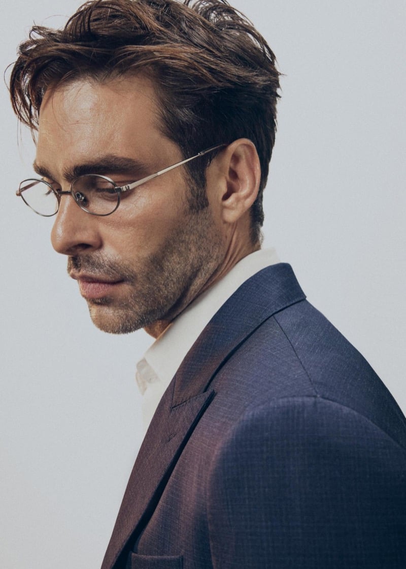 Jon wears round wire-rimmed glasses with a navy micro-check suit jacket and a cream open-collar shirt, looking down in profile