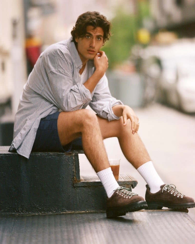 Model Tony Özkan sits on a metal stoop with an iced coffee in a striped button-up shirt, the Abercrombie A&F everyday short, white crew socks, and brown suede hiking-inspired shoes