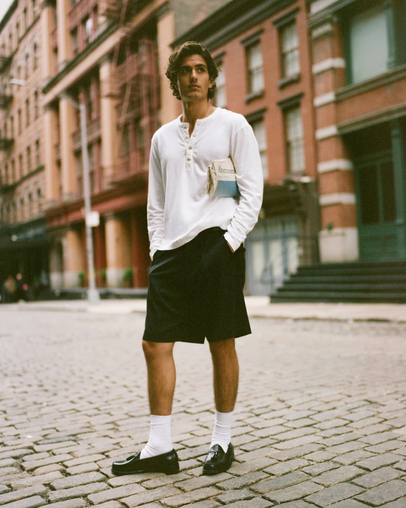 Model Tony Özkan stands on a downtown cobblestone block in a white Abercrombie & Fitch long-sleeve henley with chest patch, the brand's baggy trouser short, white crew socks, and dark leather loafers