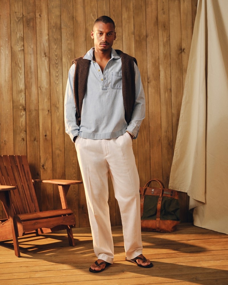 Ysham Jackson in a light-wash Abercrombie & Fitch chambray denim popover shirt with a brown crewneck sweater draped across his shoulders, white linen drawstring trousers, and black flip-flops in a wood-paneled cabin