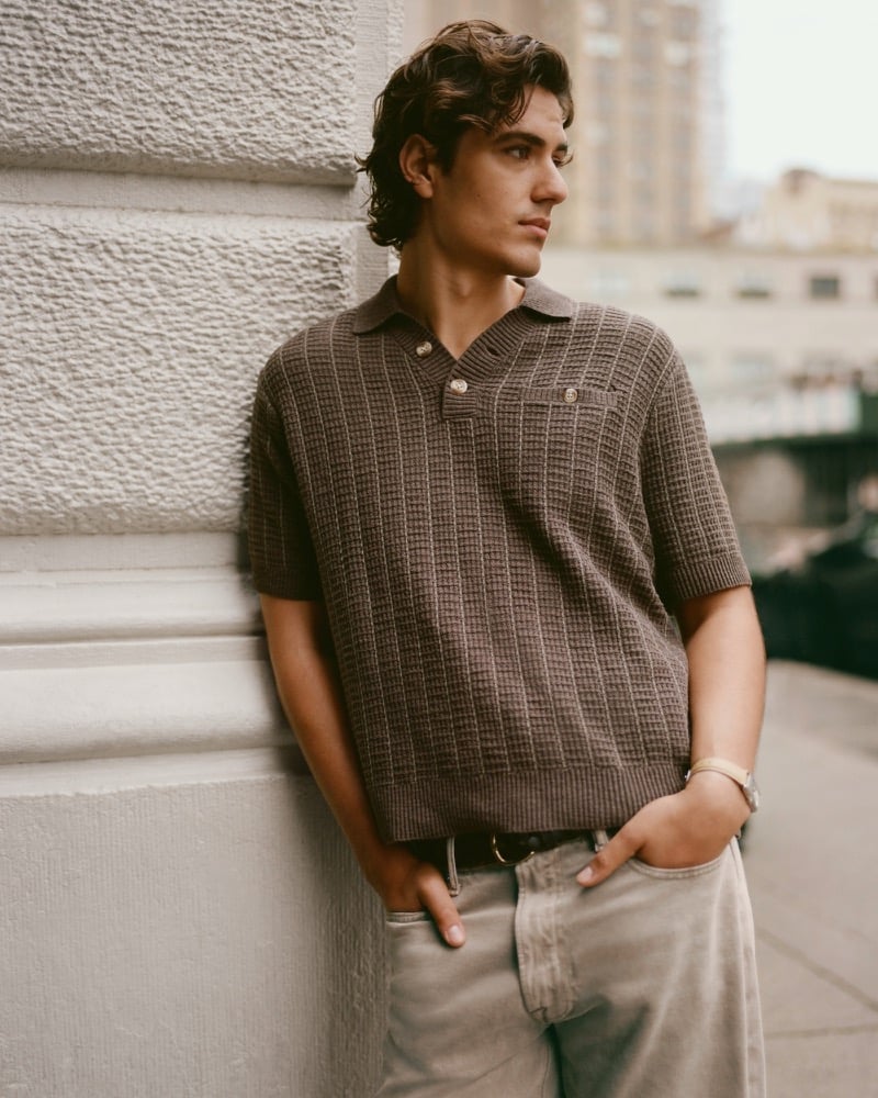 Model Tony Özkan leans against a stone facade in a brown waffle-knit Abercrombie & Fitch cotton 2-button sweater polo, tan-wash jeans, and a leather belt