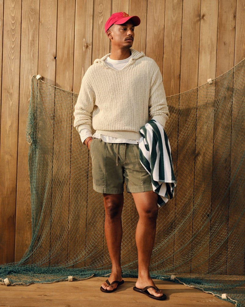 Ysham Jackson in a red Abercrombie & Fitch baseball cap, cream cotton crochet-style hooded popover sweater over a white tee, olive corduroy pull-on shorts, and flip-flops, holding a green and white striped beach towel against a fishing-net backdrop