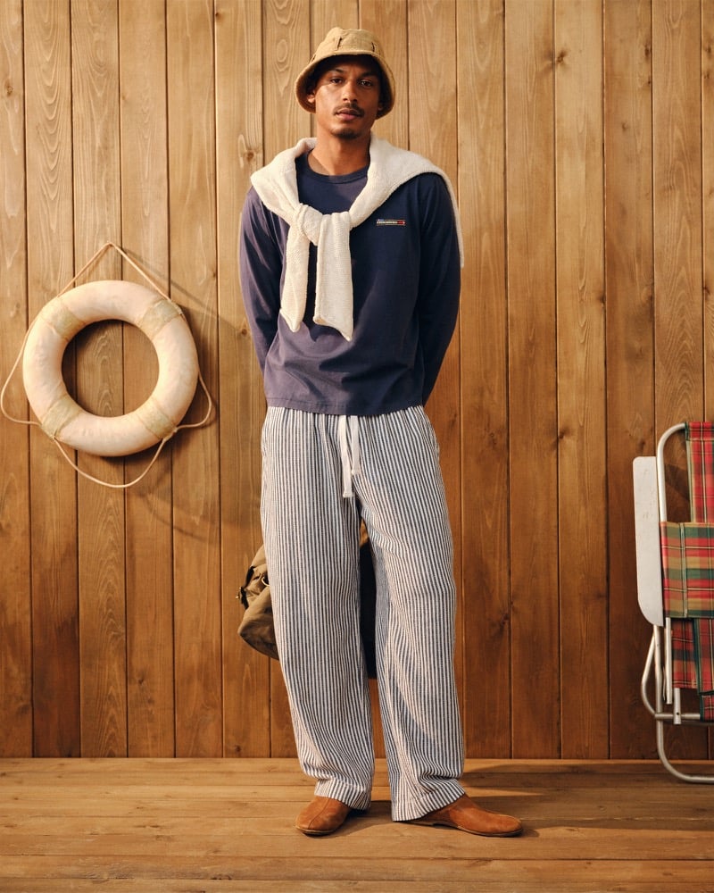 Ysham Jackson in a tan bucket hat, a navy Abercrombie & Fitch long sleeve boxy tee with a small chest patch, a cream knit sweater tied around his shoulders, blue and white striped baggy pull-on pants, and brown suede loafers