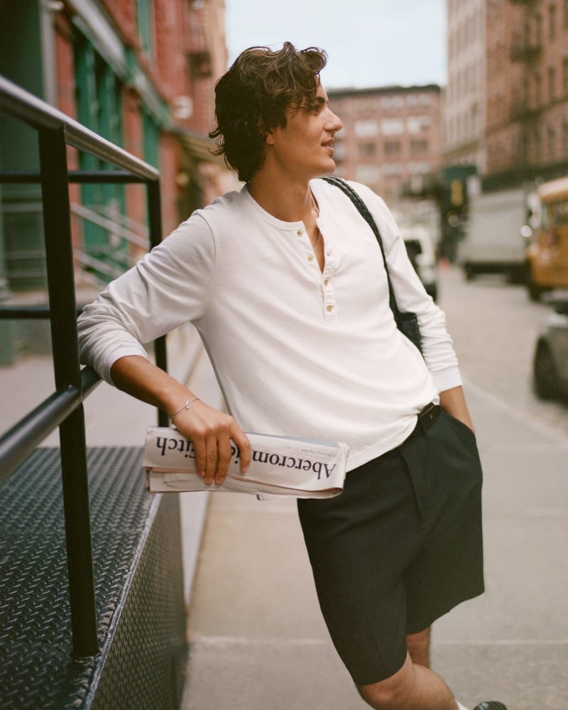 Model Tony Özkan leans on a metal stoop railing in the Abercrombie & Fitch long-sleeve linen-blend henley with dark pleated shorts, holding a folded Abercrombie & Fitch newspaper