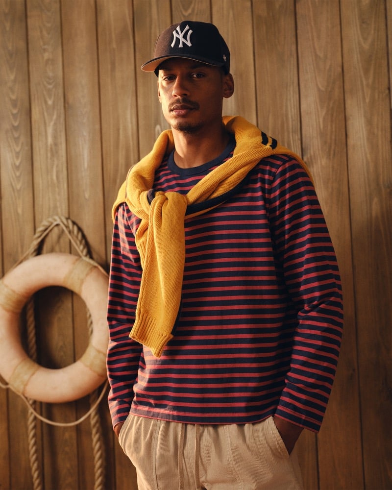 Ysham Jackson in a navy New York Yankees cap and a navy and red striped Abercrombie & Fitch long sleeve boxy tee with a mustard yellow knit sweater knotted around his shoulders, khaki trousers below, posed beside a hanging life ring