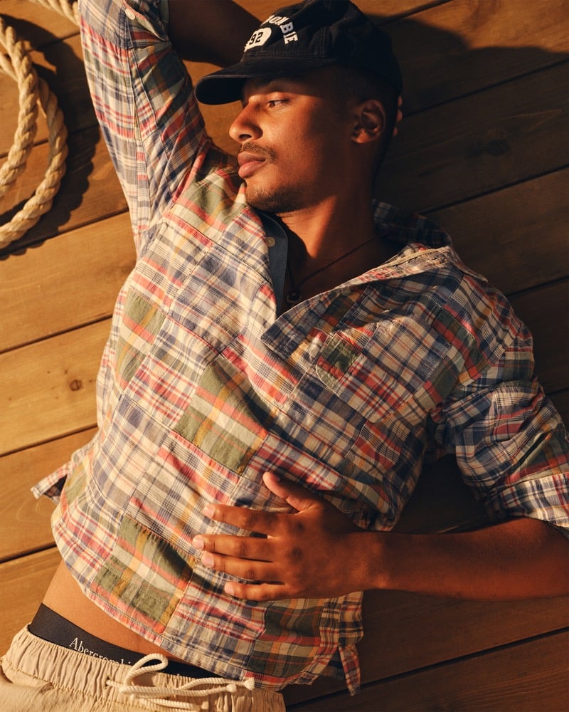 Ysham Jackson lying back on a wood-planked deck in a navy Abercrombie & Fitch baseball cap and a patchwork madras popover shirt in red, blue, and green checks, with thick rope coiled beside him