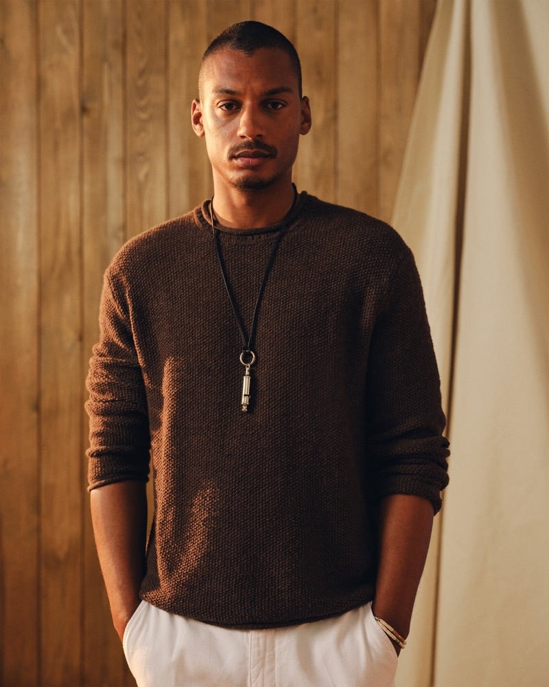 Portrait of Ysham Jackson in a chocolate brown Abercrombie & Fitch cotton rollneck crew sweater paired with white pull-on trousers, a long silver whistle pendant resting at his chest