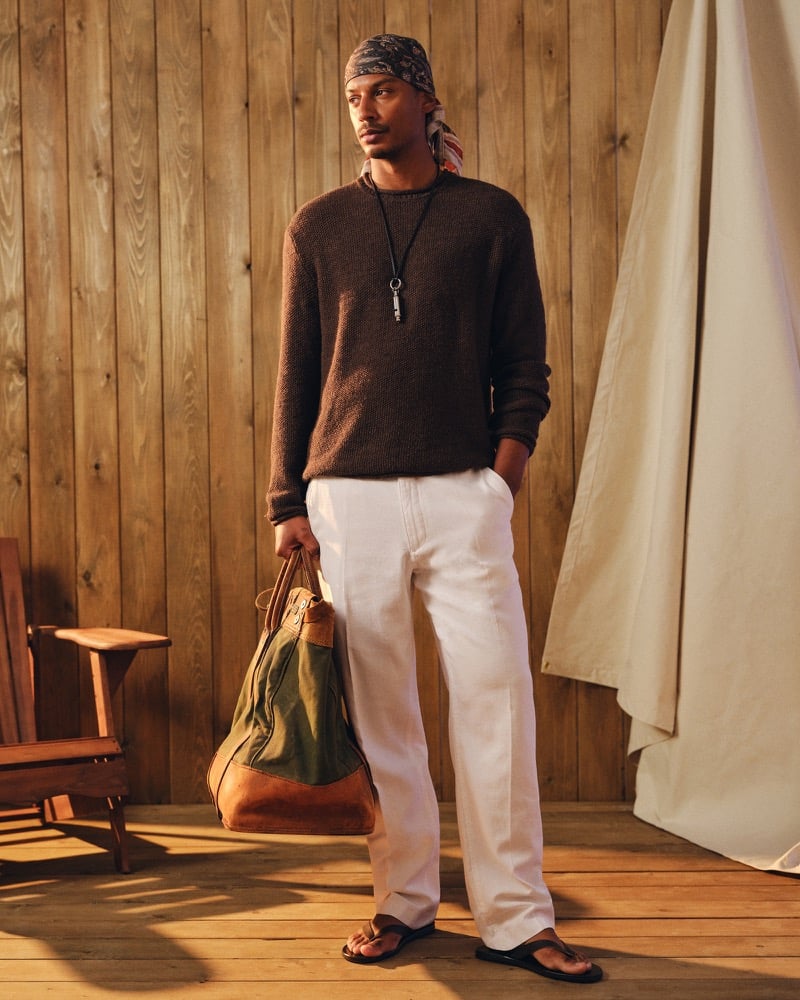 Ysham Jackson in a patterned bandana headscarf, a brown cotton crew sweater, white pull-on trousers, and brown flip-flops, carrying an olive and tan canvas weekender bag for Abercrombie & Fitch