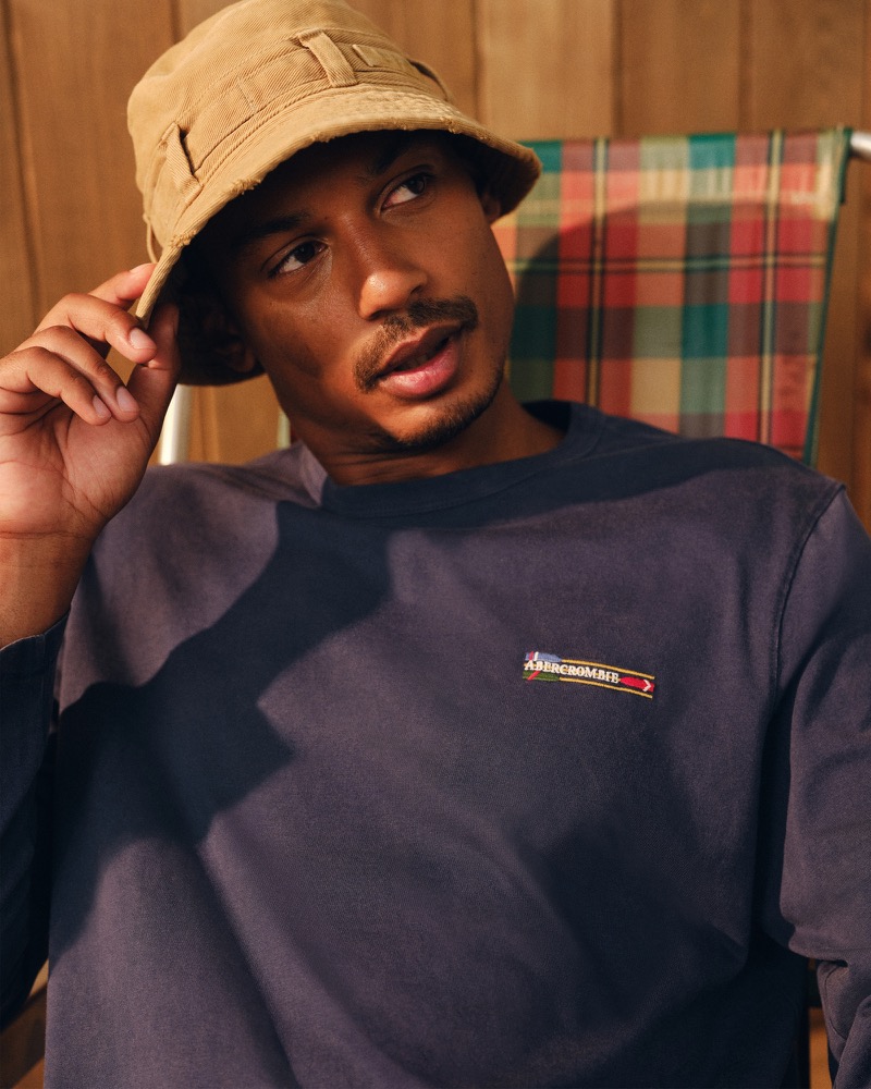 Ysham Jackson in a tan canvas bucket hat and a faded navy Abercrombie & Fitch crewneck sweatshirt with a small embroidered logo patch, seated on a plaid camp chair