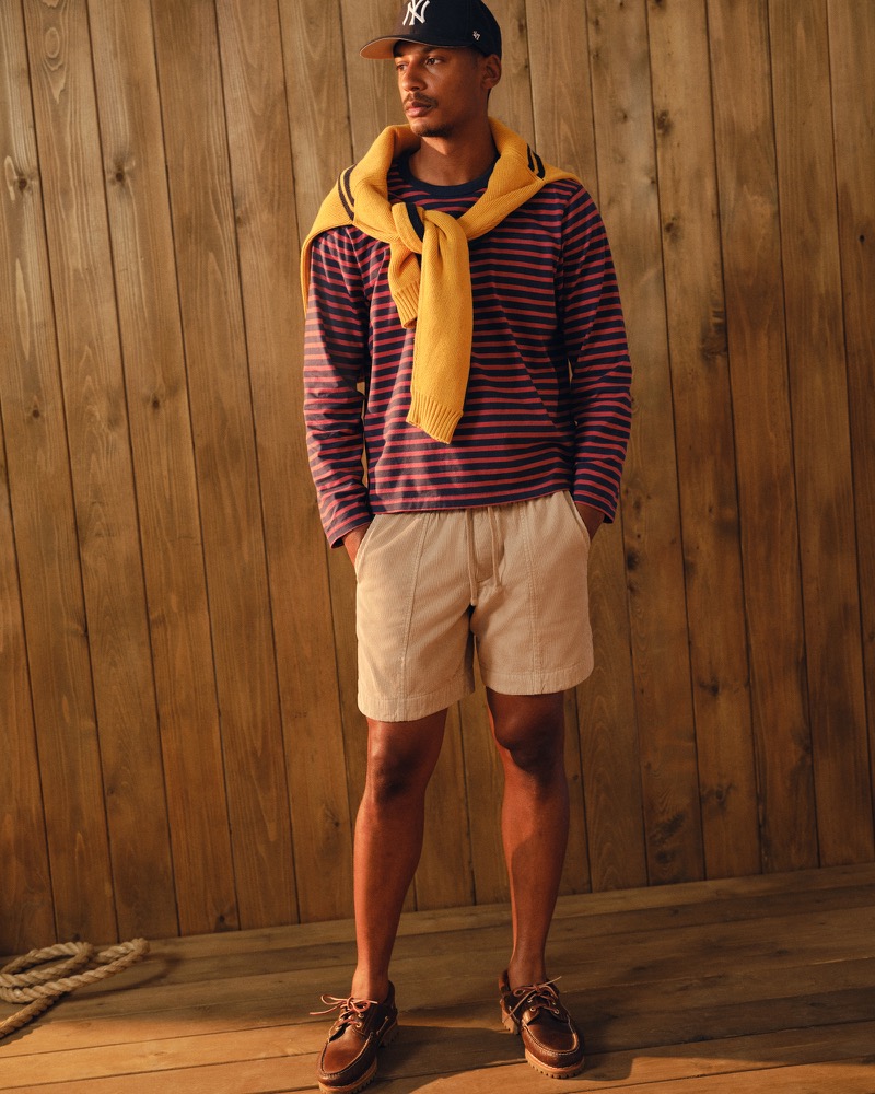 Ysham Jackson in a New York Yankees cap and a navy and red striped Abercrombie & Fitch long sleeve boxy tee with a mustard knit knotted at the shoulders, khaki shorts, and brown boat shoes