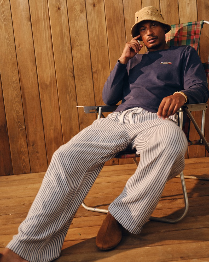 Ysham Jackson seated in a metal-framed camp chair in a navy Abercrombie & Fitch crewneck sweatshirt, blue and white striped baggy pull-on pants, and brown suede loafers