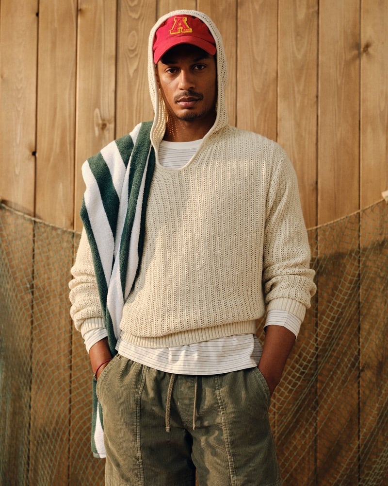 Ysham Jackson in a red Abercrombie & Fitch baseball cap and cream crochet-style hooded popover with a green and white striped beach towel slung across the shoulder, olive corduroy shorts below