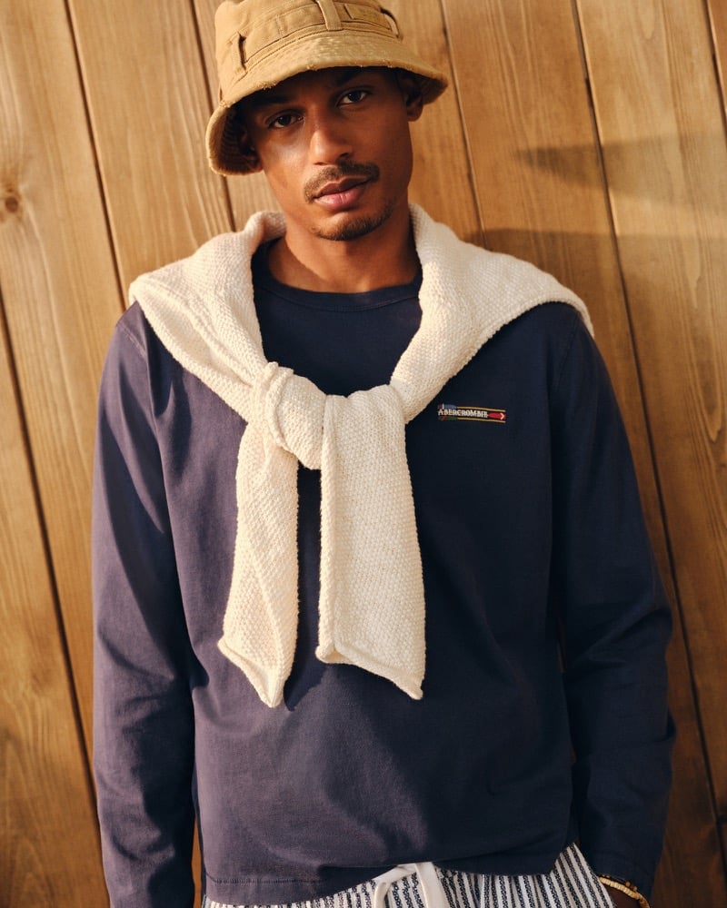 Ysham Jackson in a tan bucket hat and a navy Abercrombie & Fitch long sleeve tee with a small striped chest patch, a cream textured knit sweater tied around his shoulders