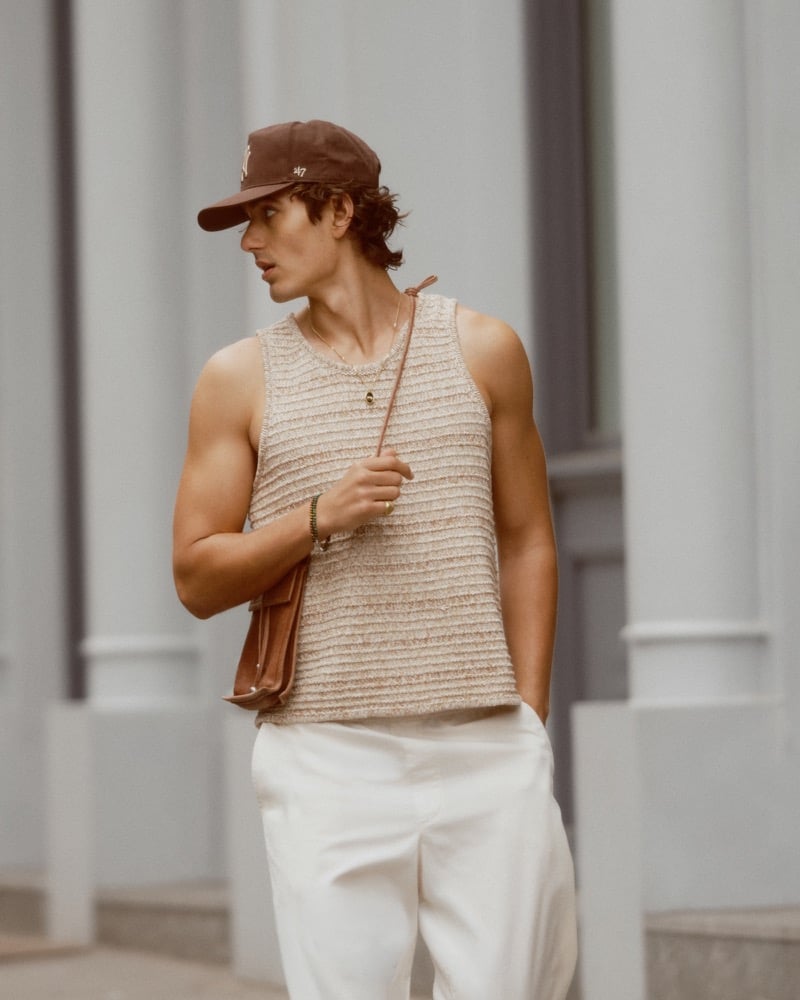 Model Tony Özkan wears the Abercrombie & Fitch textural sweater tank with pale pull-on barrel pants, a brown 47 baseball cap, and a brown leather crossbody bag in front of a SoHo storefront