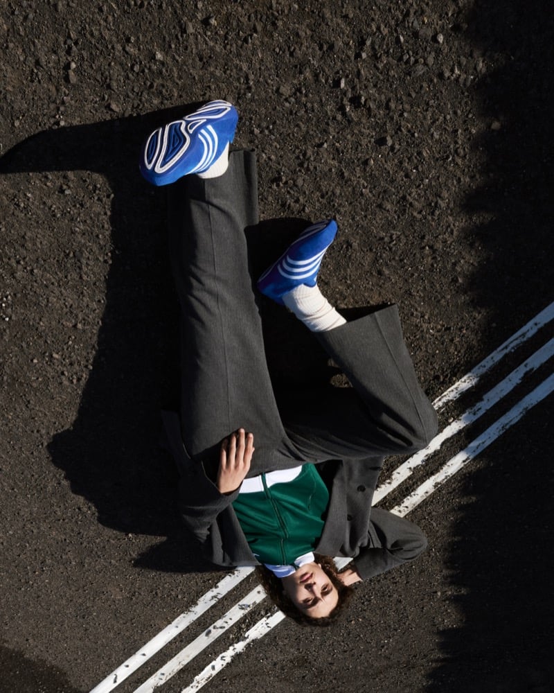 Overhead view of Mohamed Benhadda lying on earth in a gray wool suit, green track jacket, white tube socks, and blue suede Adidas Originals Pharrell Williams Virginia Vario Flat Earther sneakers