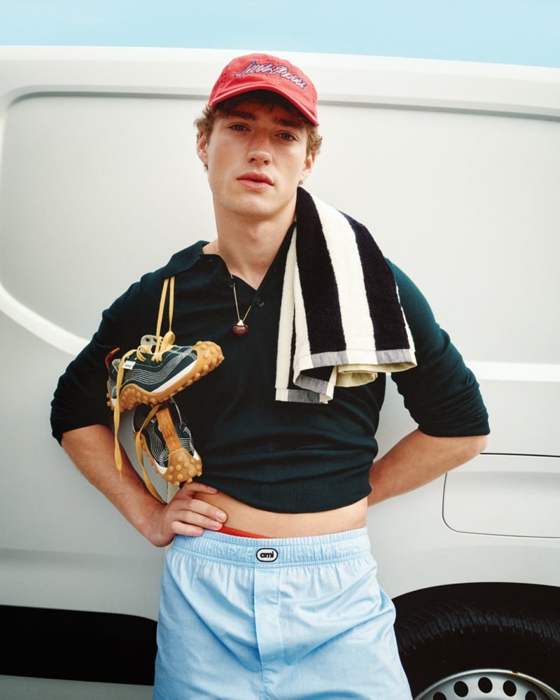 Valentin Humbroich wears a red Ami Paris cap with a long-sleeve polo and light blue Ami-logo boxer shorts while holding a pair of Mirage sneakers and a striped towel over his shoulder