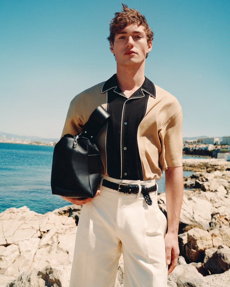 Valentin Humbroich wears a tan and black colorblock Ami Paris bowling shirt with cream pleated trousers and a braided leather belt, holding a black leather bucket bag against a Mediterranean coastline