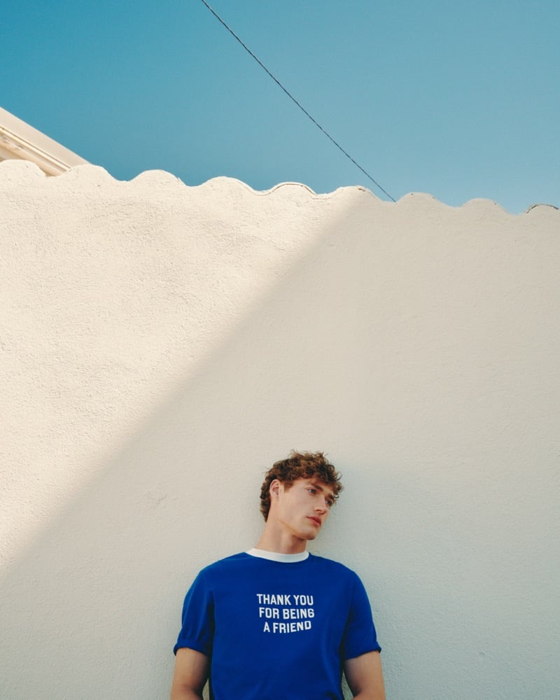 Valentin Humbroich stands against a sunlit white stucco wall in the royal blue Ami Paris "Thank You For Being a Friend" t-shirt under a bright Mediterranean sky