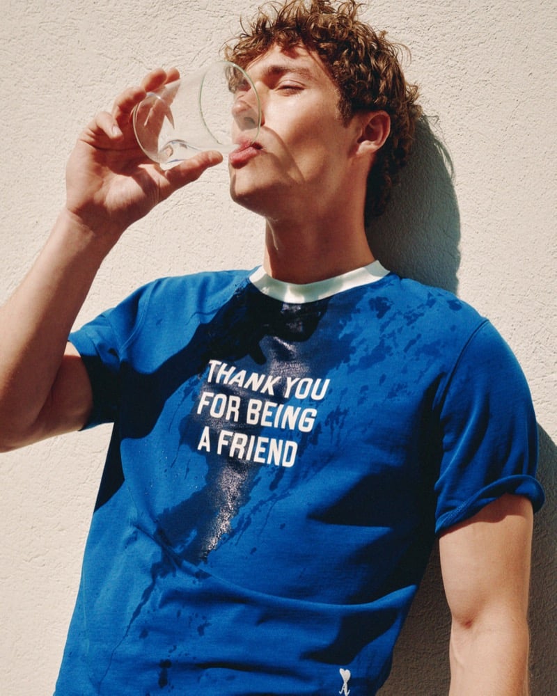 Valentin Humbroich sips from a glass of water against a sunlit wall wearing a royal blue Ami Paris "Thank You For Being a Friend" t-shirt from the 15th anniversary capsule