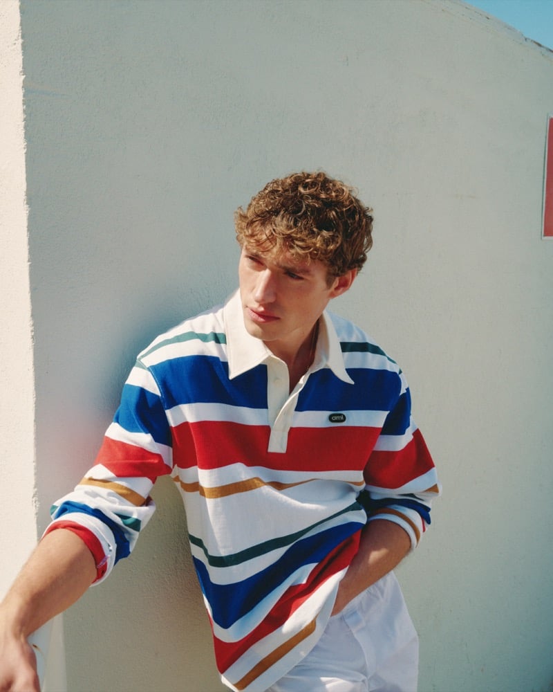 Valentin Humbroich leans against a pale blue wall in an Ami Paris striped rugby polo in red, blue, white, mustard, and green with white trousers