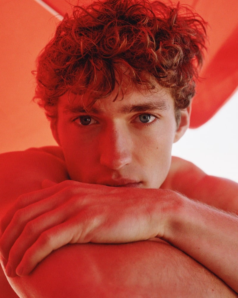 Close-up of Valentin Humbroich resting his chin on his folded arms beneath an orange beach parasol that bathes the frame in warm light