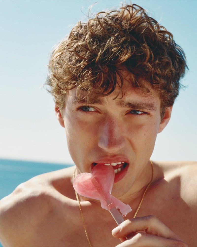 Close-up of Valentin Humbroich biting into a pink ice pop with a thin gold chain visible at his collarbone for the Ami d'été campaign