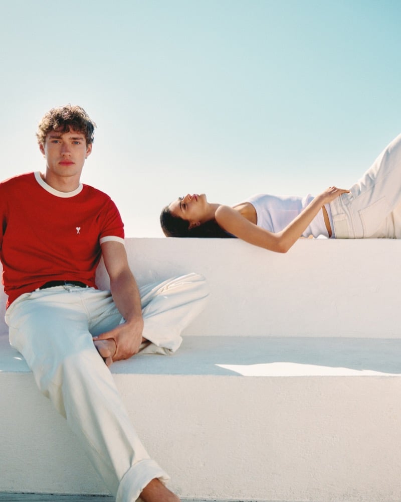Valentin Humbroich sits cross-legged on a whitewashed ledge in a red Ami de Coeur ringer t-shirt and off-white trousers beside a woman reclining in a white tank and cream trousers