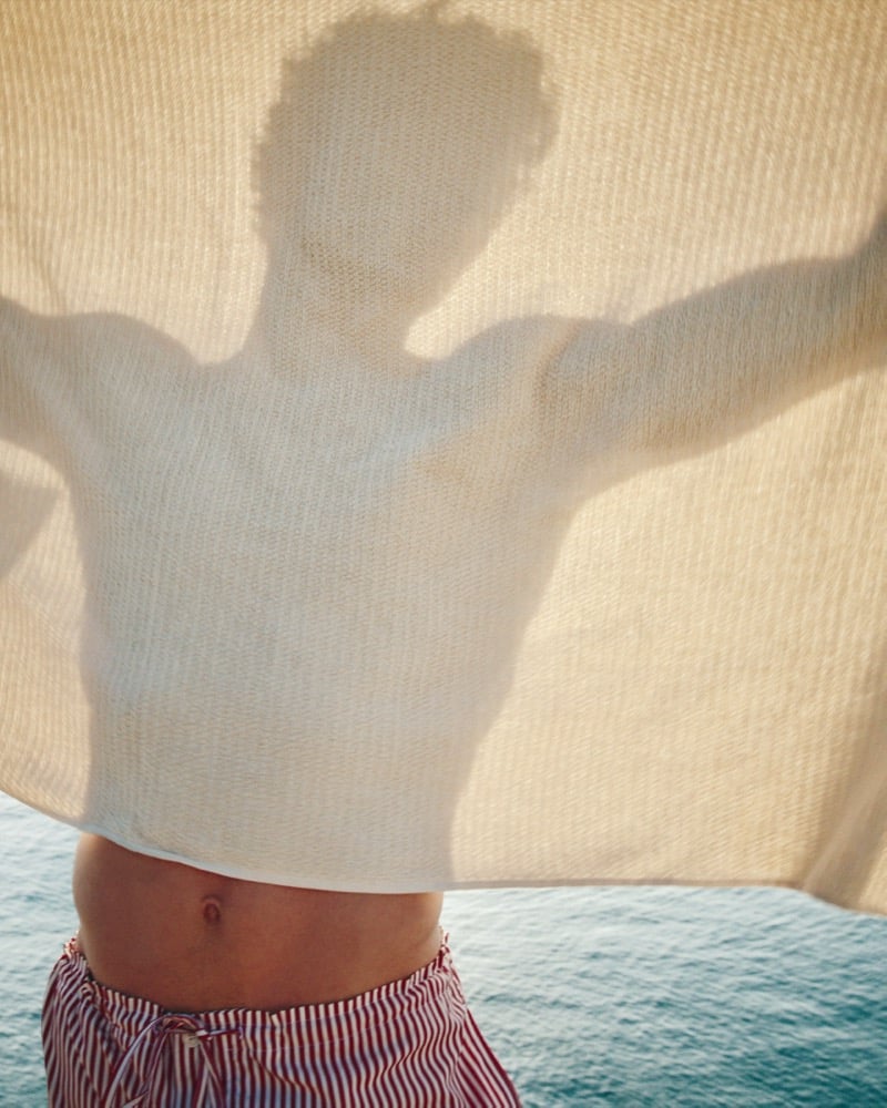 Silhouette of Valentin Humbroich pressed against a cream knit beach blanket while wearing red and white striped Ami Paris swim shorts on the water