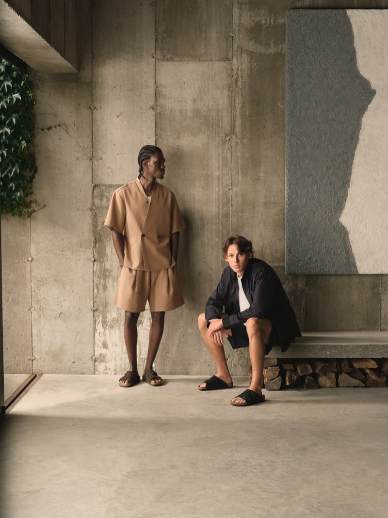 Anarcius Jean wears a camel short-sleeve buttoned shirt with matching shorts and woven brown sandals beside Samuel W. in a shirt jacket, white tee, shorts, and black slides against a concrete wall