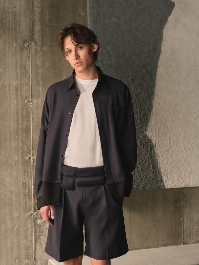 Samuel W. wears a charcoal overshirt with black leather collar and cuffs over a white tee, paired with matching charcoal pleated shorts