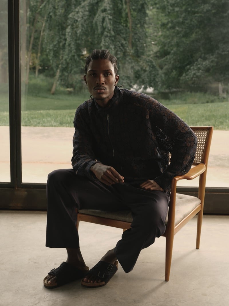 Anarcius Jean sits in a cane-back chair in a black floral lace zip-front mock-neck shirt with black trousers and black Birkenstock slides