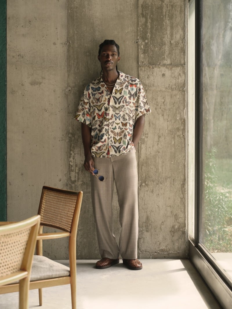 Anarcius Jean wears a butterfly print short-sleeve camp shirt with beige windowpane wide-leg trousers and woven brown loafers, holding round blue sunglasses