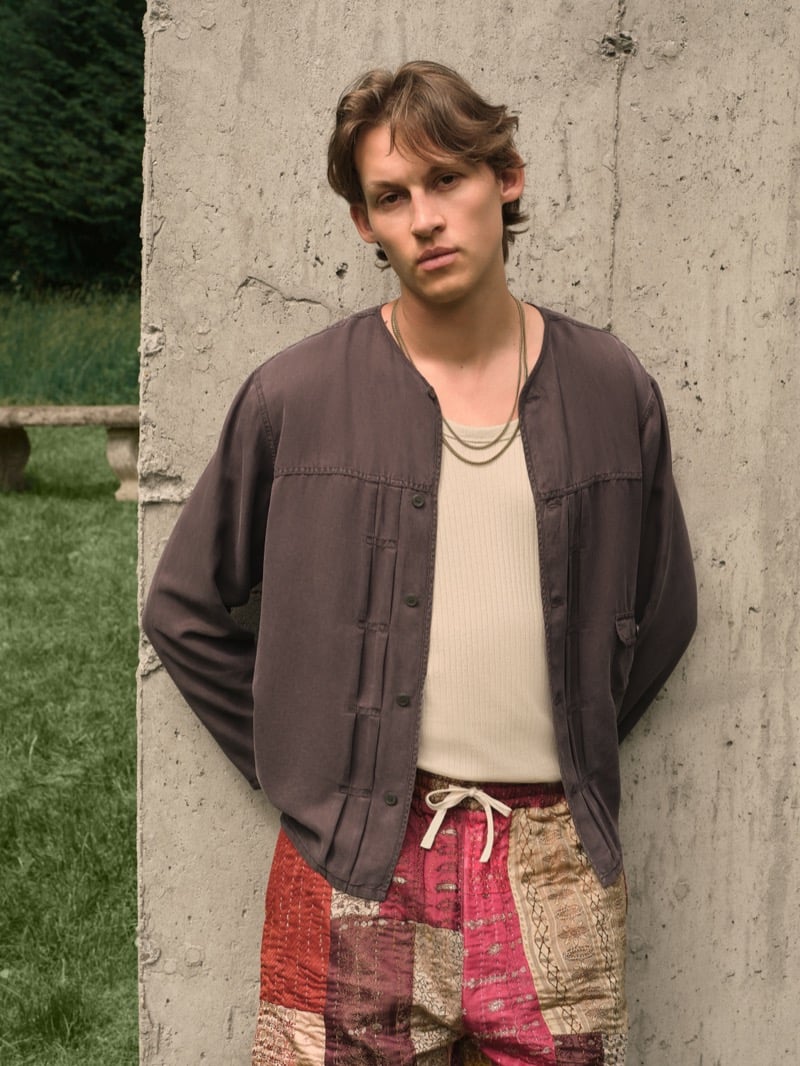 Samuel W. wears a washed gray collarless jacket over a beige ribbed tank with red, pink, and gold patchwork drawstring shorts against a concrete wall