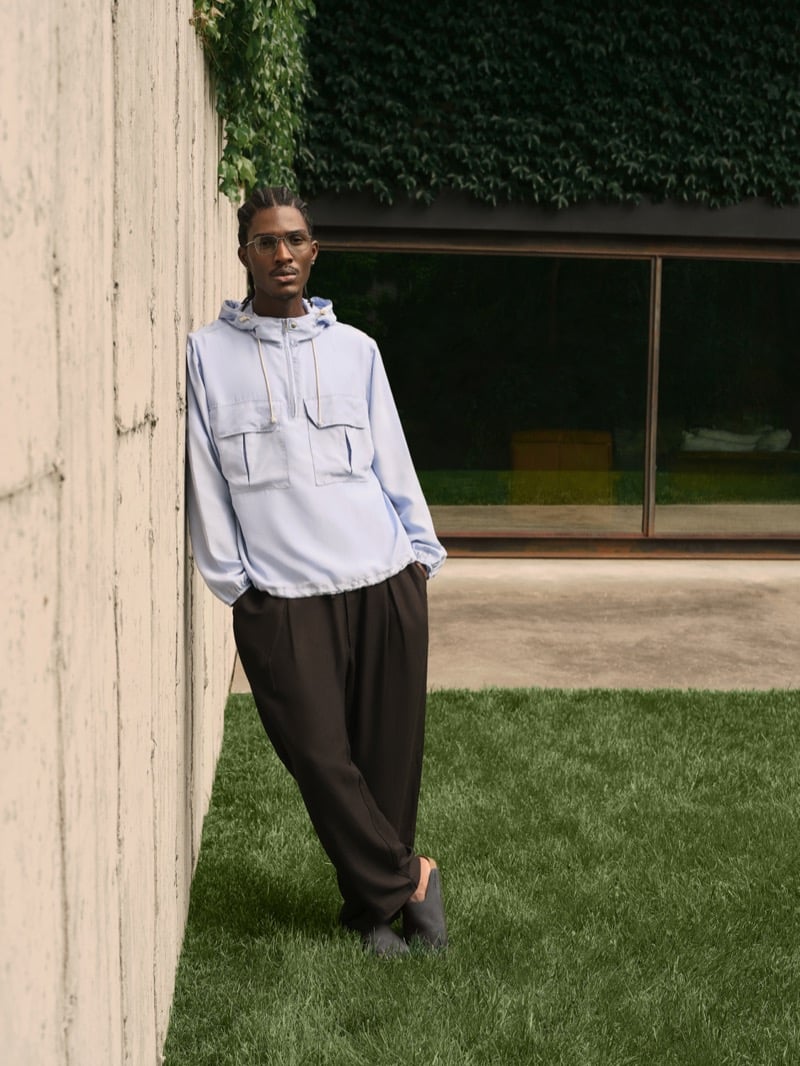 Anarcius Jean wears wire-rim glasses with a pale blue hooded anorak with kangaroo pockets, dark brown wide-leg pleated trousers, and gray slides on a lawn