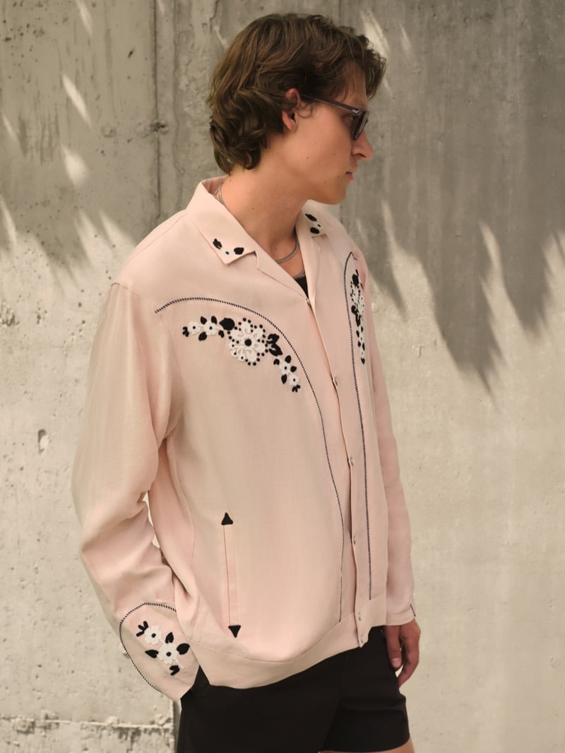 Samuel W. wears tortoiseshell glasses and a blush pink western-inspired camp shirt with black and white floral embroidery and contrast stitching, paired with black shorts