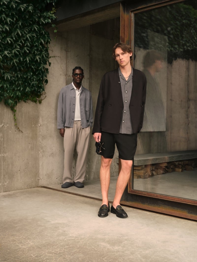 Anarcius Jean stands in dark sunglasses with a gray linen overshirt and beige pleated trousers, while Samuel W. stands in front in a black collarless blazer, gray button-up, black shorts, and black bit loafers