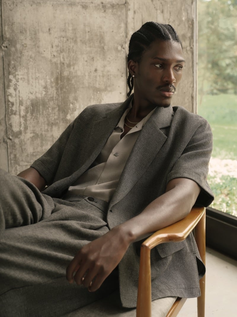 Anarcius Jean leans against a concrete wall in a gray short-sleeve linen blazer with matching pleated trousers, an olive shirt, and layered gold chains