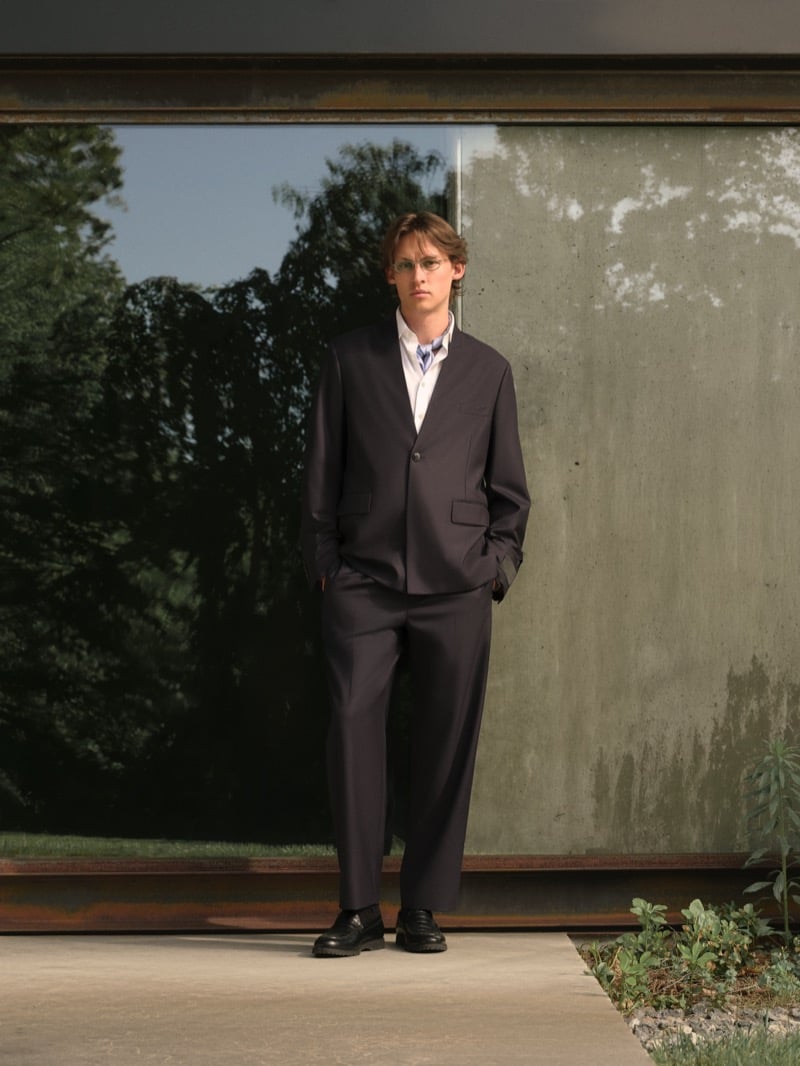 Samuel W. wears round wire-rim glasses with a collarless single-breasted suit, white button-up shirt, and black lug-sole derbies