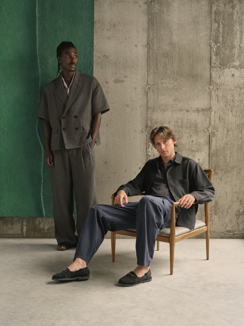 Anarcius Jean stands in a gray double-breasted short-sleeve blazer with matching wide-leg trousers and brown sandals beside Samuel W., who sits in a black overshirt, gray polo, navy seersucker pleated trousers, and black woven loafers