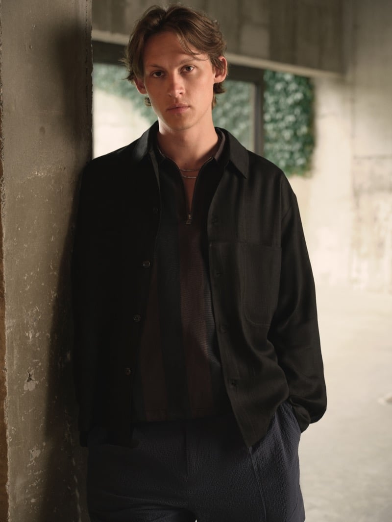 Samuel W. wears a black overshirt over a black zip mock-neck knit with dark gray seersucker pleated trousers against a concrete pillar