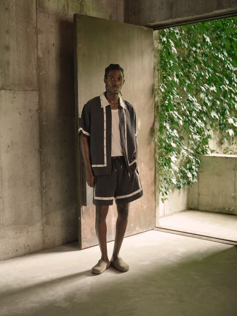 Anarcius Jean wears a black camp collar shirt with cream piping and graphic trim, a white ribbed tank, matching black drawstring shorts, and beige clog-style slides in a concrete doorway