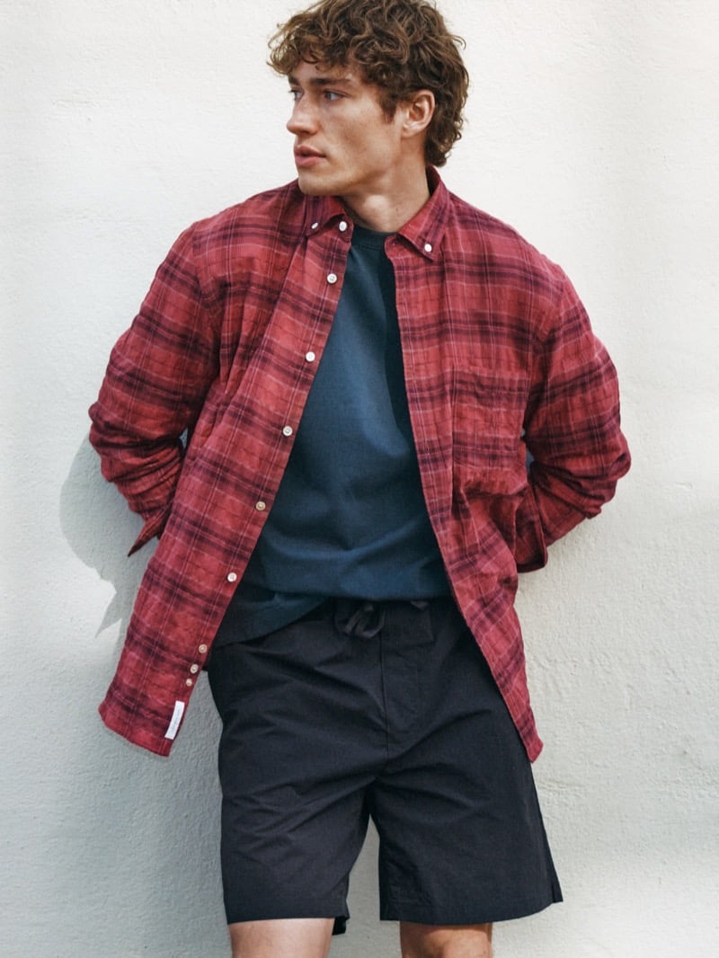 Valentin Humbroich wears an open washed red plaid button-down over a dark T-shirt with dark drawstring shorts against a concrete wall