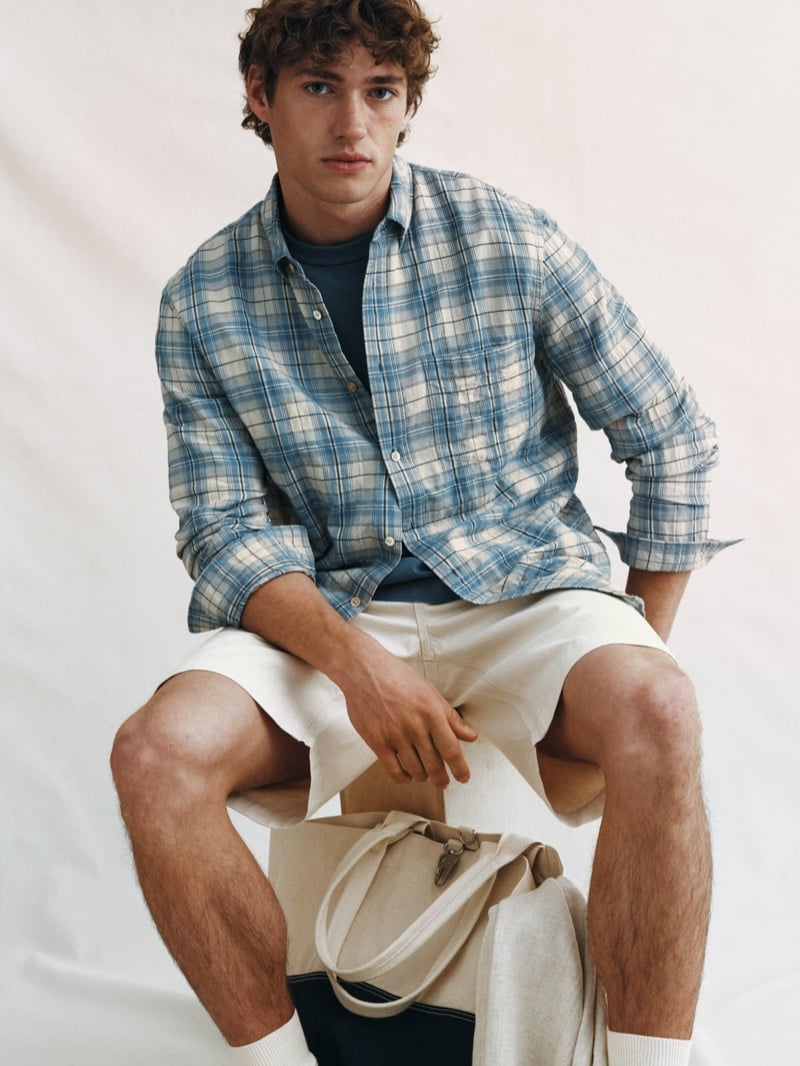 Valentin Humbroich wears a sun-faded blue plaid button-down over a dark T-shirt with ecru cotton shorts, perched on a stool with a canvas tote